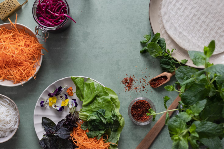 Various fresh ingredients such as shredded carrots, purple cabbage, lettuce, herbs, and rice noodles, arranged on a green surface.